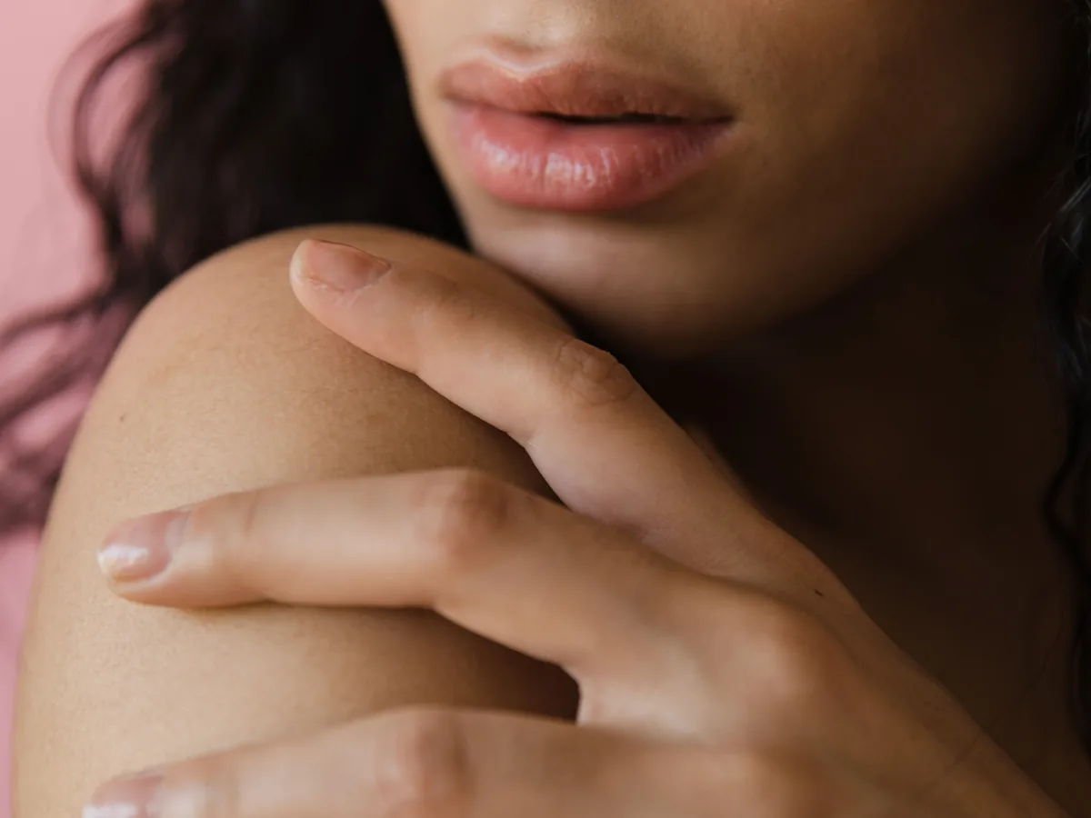 Close up of a woman's hand resting gently on her bare shoulder, natural skin against a warm pink backdrop